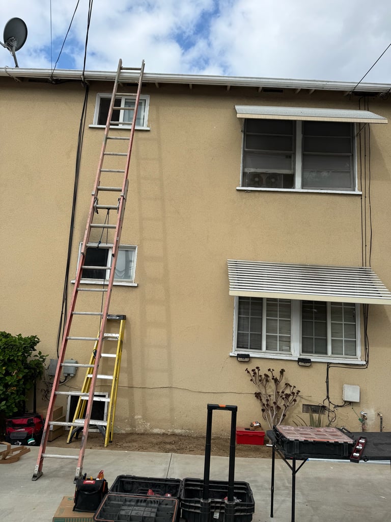 Tall metal ladder leaning against tan-colored house exterior with windows, outdoor work setup visible below