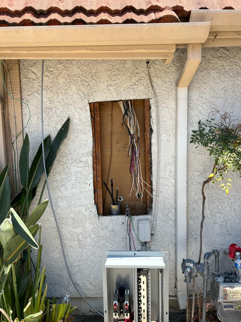 Exterior electrical panel and junction box on stucco wall with hanging wires, potted plants, and pergola overhead