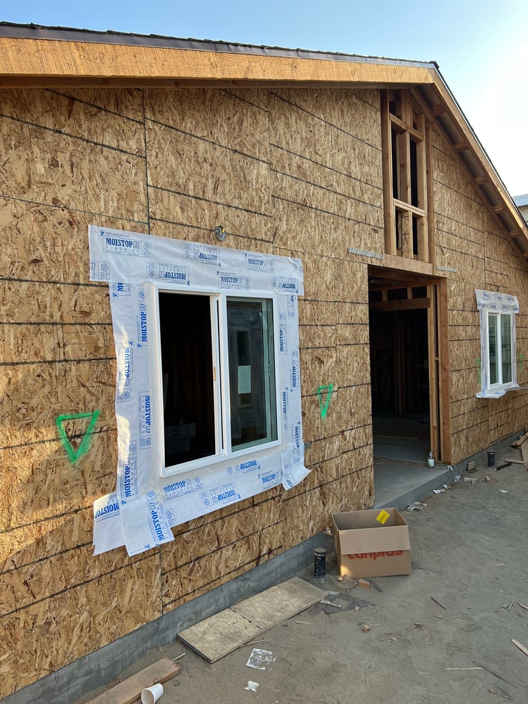 House under construction with OSB sheathing, weatherproof wrap around window opening, and blue building paper covering