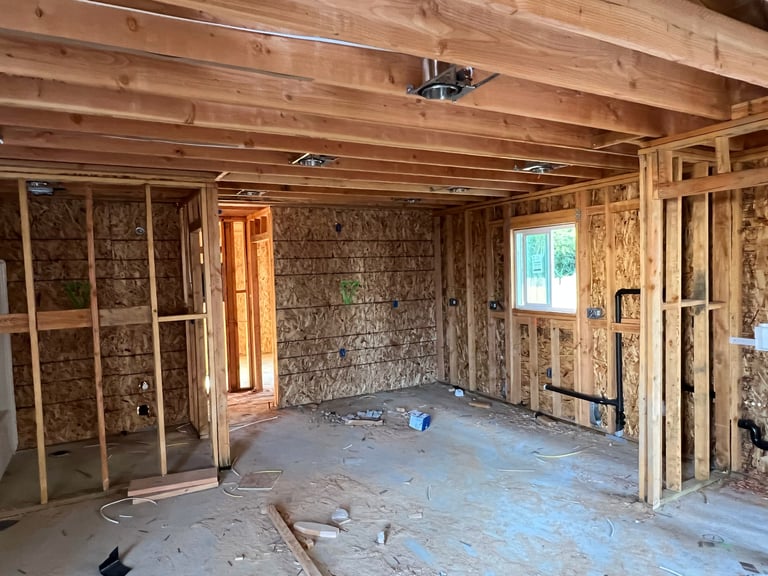 Interior of a home under construction showing exposed wooden framing, insulation, and concrete floor with various construction materials and tools scattered around