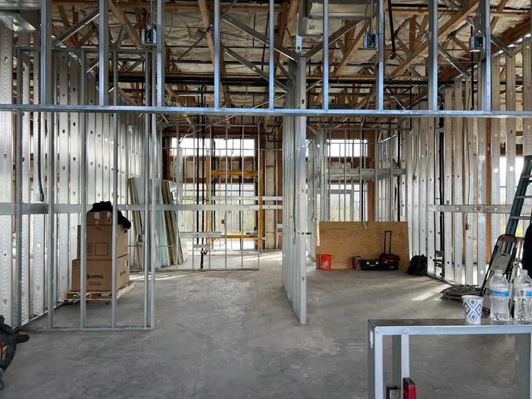 Interior of a residential construction site showing metal framing, drywall, exposed studs, and construction materials on a concrete floor