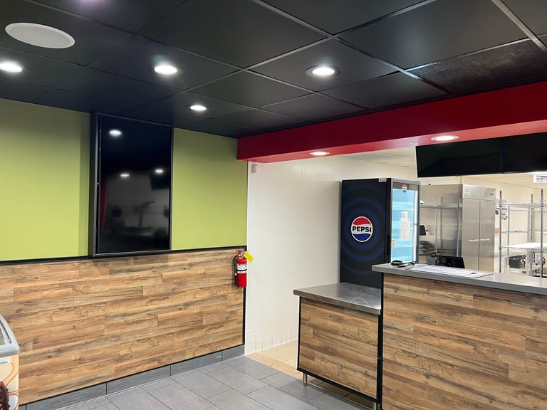 Interior of a modern fast-casual restaurant counter area with lime green and burgundy walls, wooden countertops, and bright ceiling lights