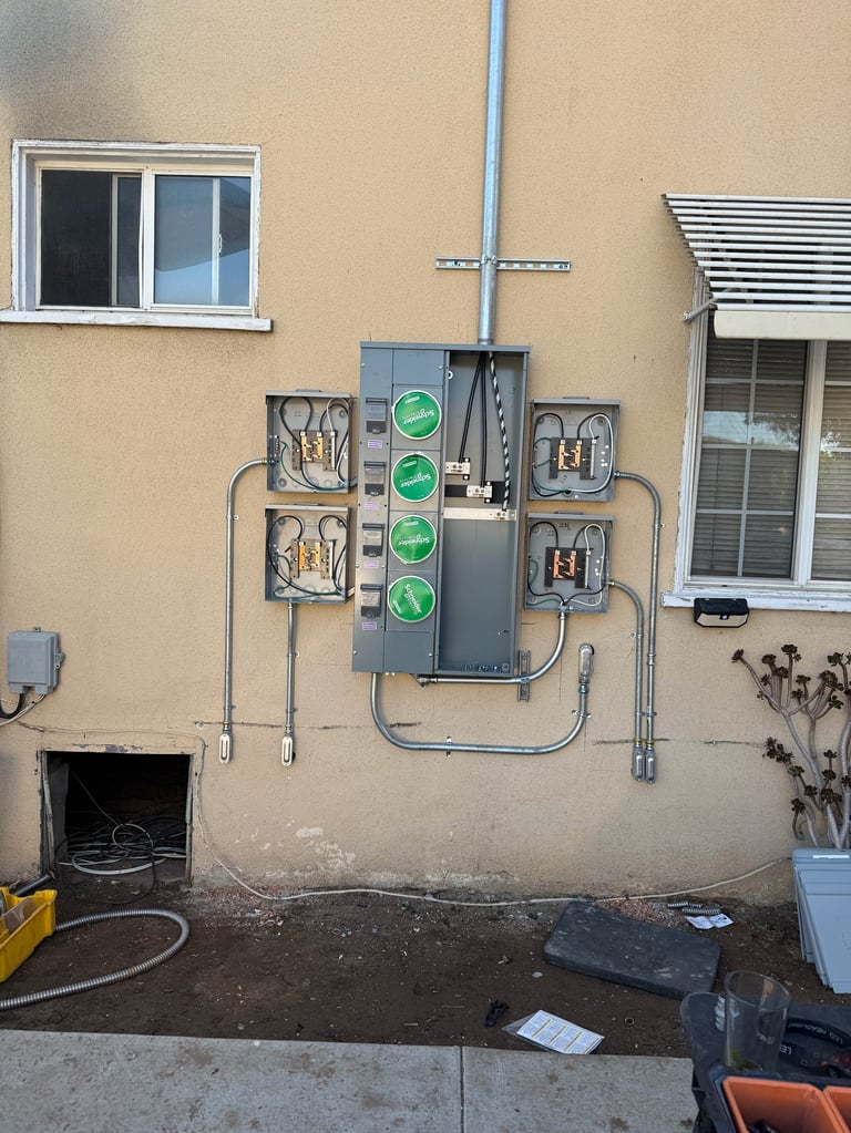 Electrical panel mounted on beige building wall with green indicator lights and metal conduit pipes, flanked by two windows