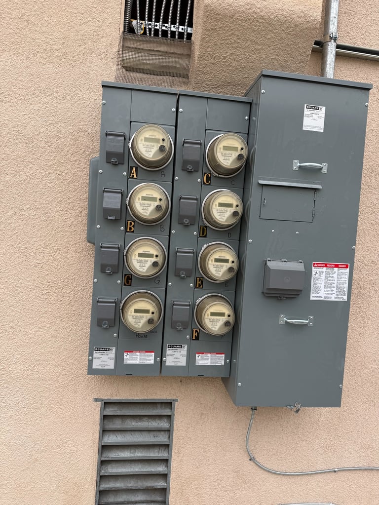 Electrical meter panel mounted on tan wall with eight round utility meters and gray switch boxes