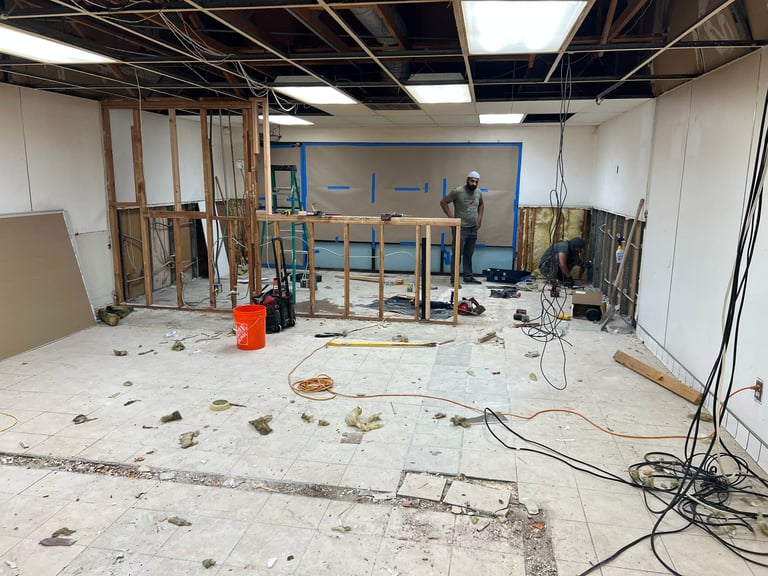 Construction site interior with exposed ceiling, wooden framing, debris scattered on floor, and electrical cords