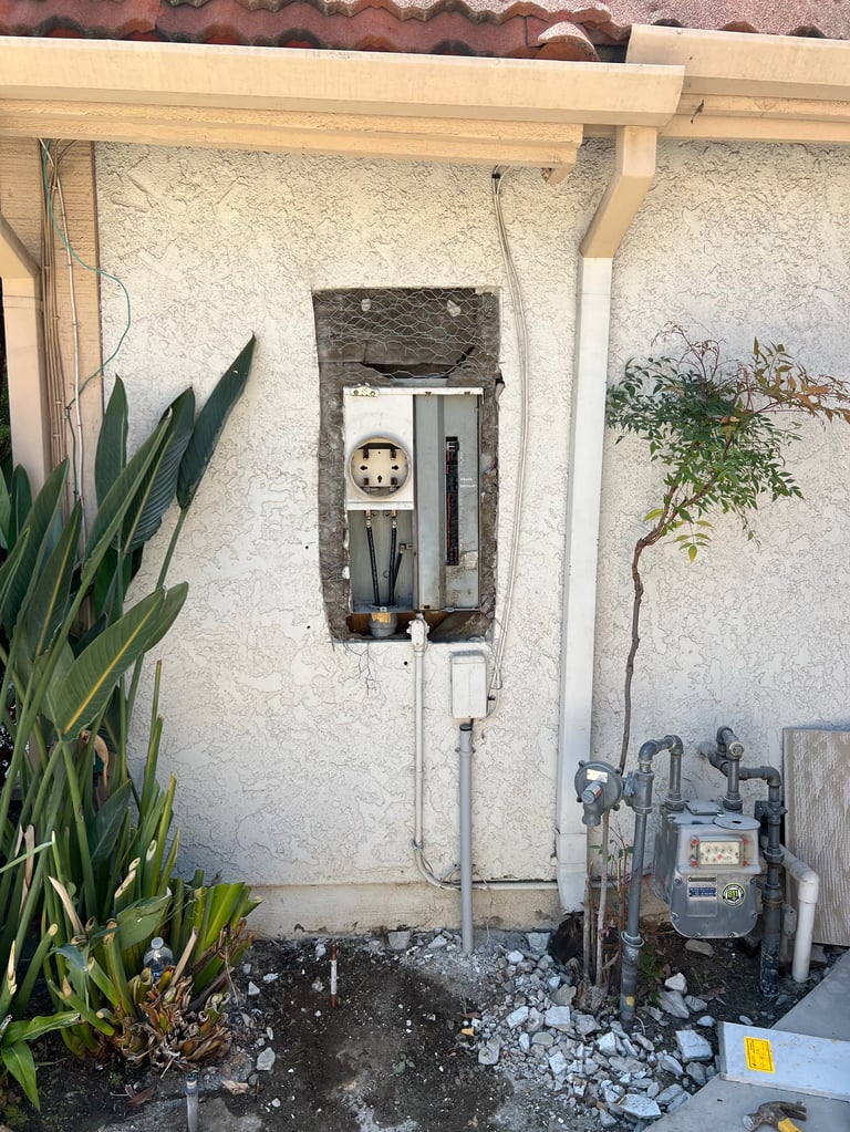 Weathered concrete wall with open electrical panel, surrounding plants, and utility meter box beneath wooden pergola