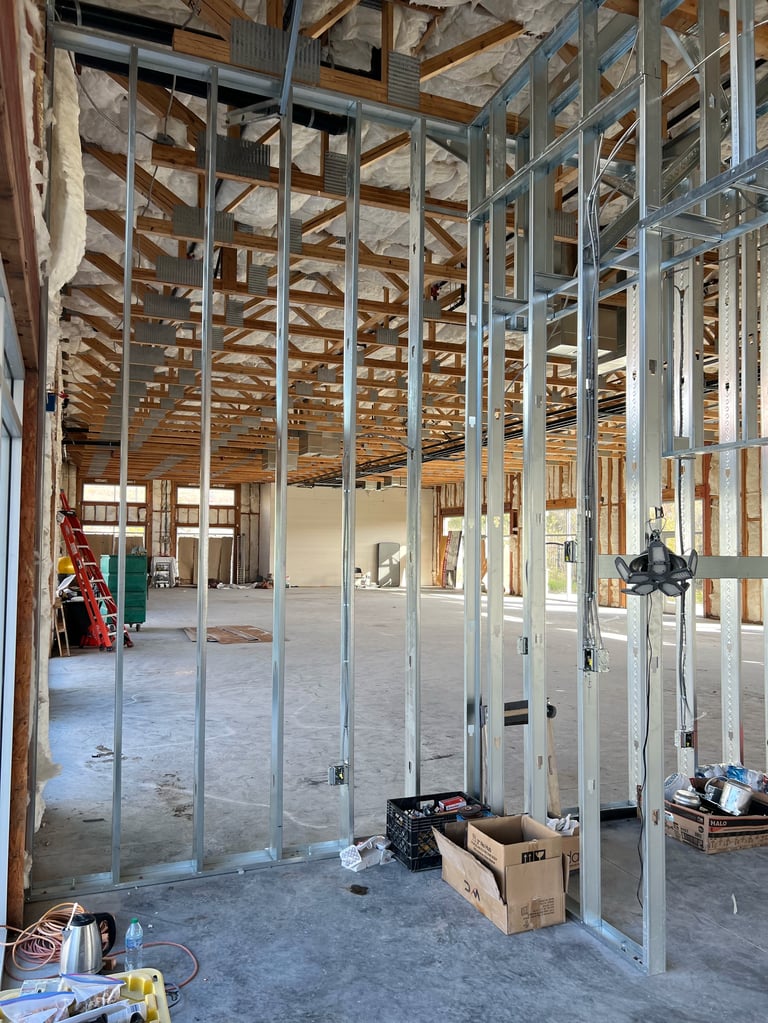 Interior of a construction site showing metal framing studs, wooden ceiling joists, concrete floor, and building materials in boxes