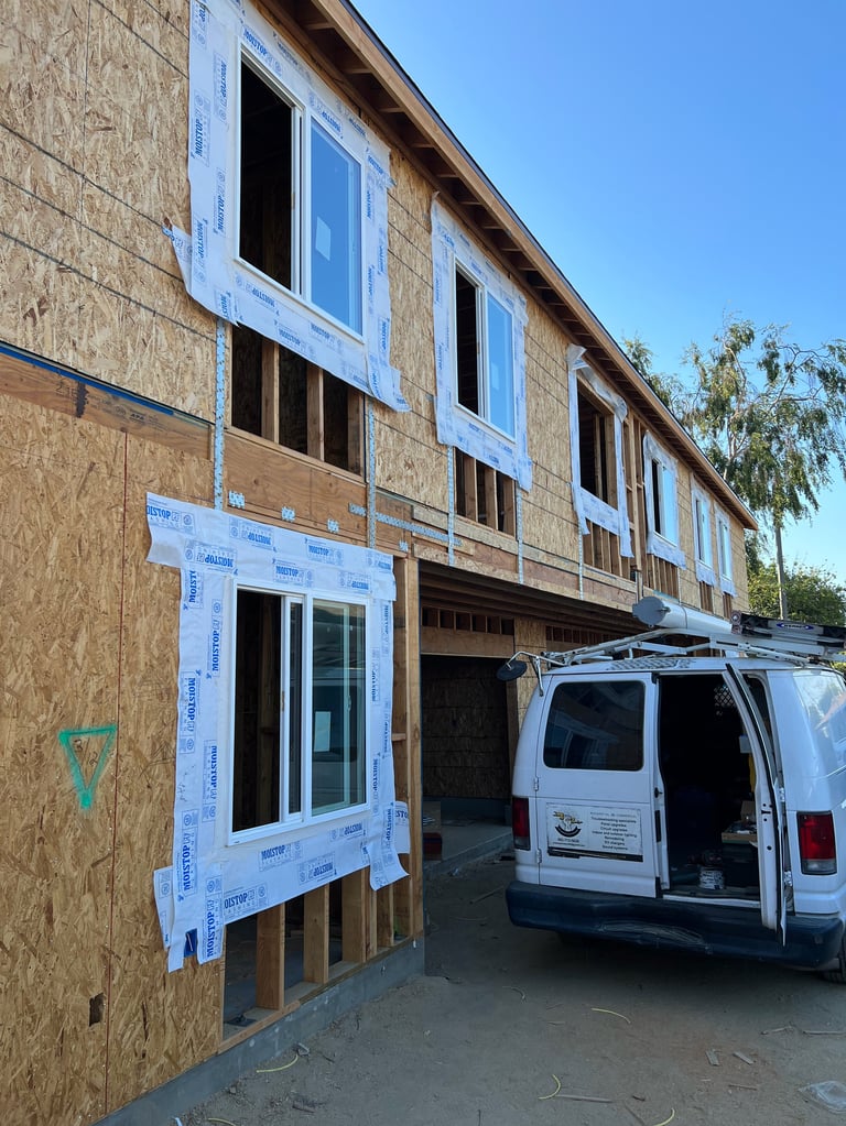 Construction site showing building exterior with OSB sheathing, blue house wrap, open window frames, and white work van parked outside