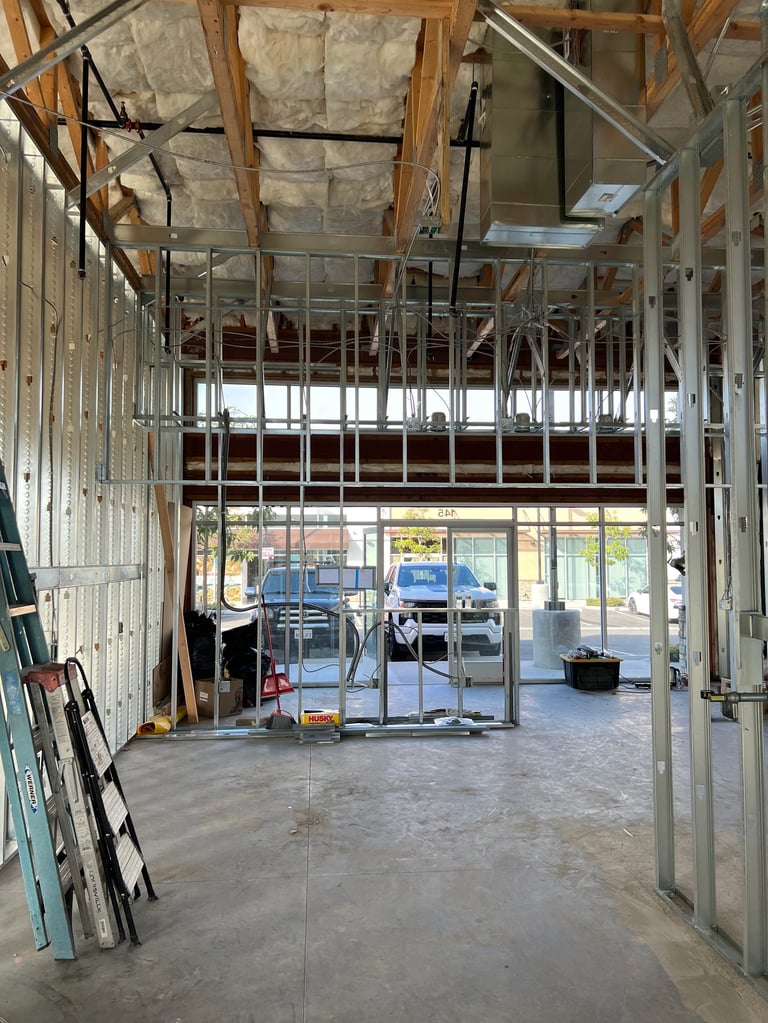 Interior of a commercial building under construction with exposed framing, concrete floor, and storefront windows visible at the entrance