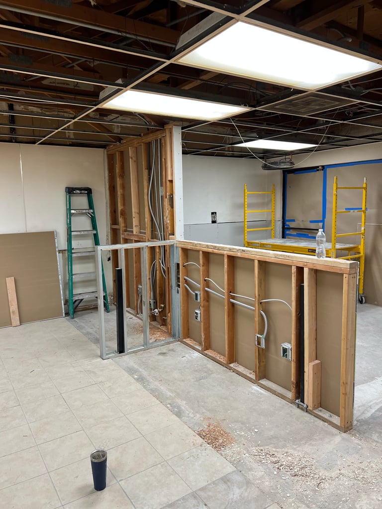 Interior construction site with wooden framing, white ceiling panels, and yellow and blue ladders against blue wall
