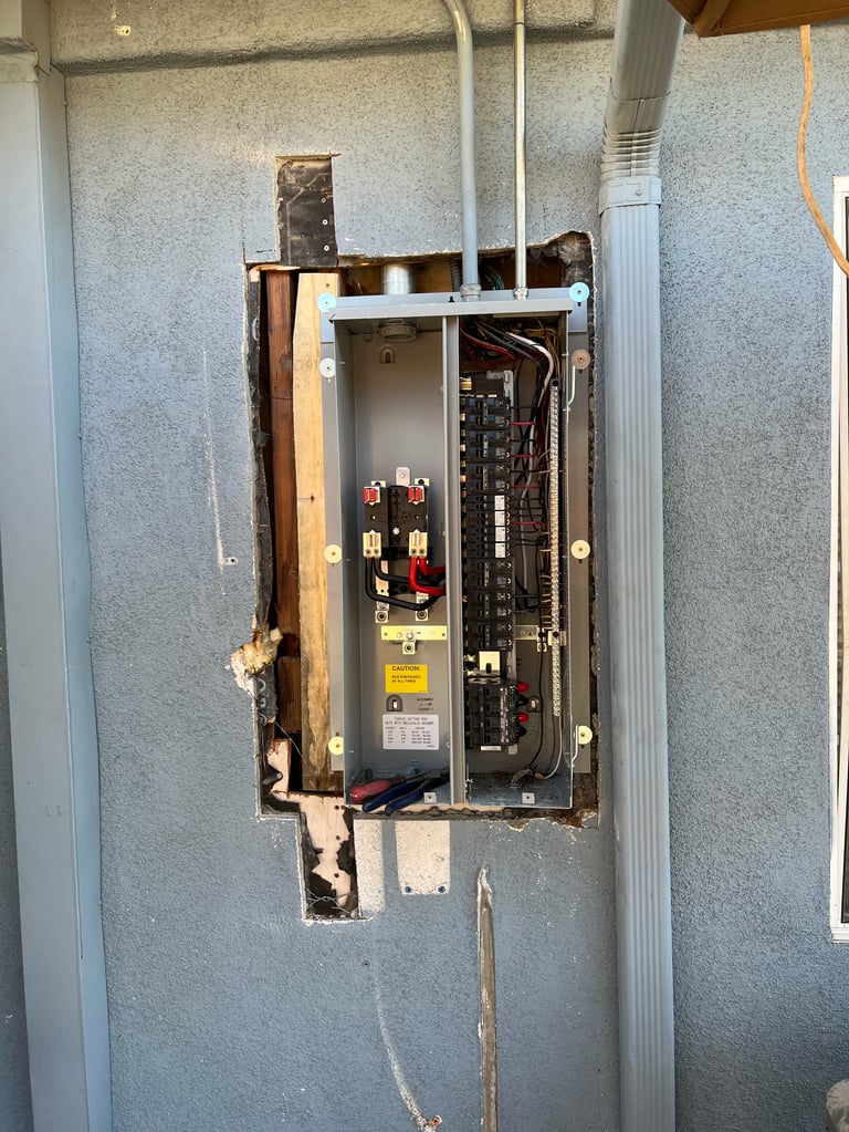 Open electrical panel box mounted on gray wall showing circuit breakers and wiring connections