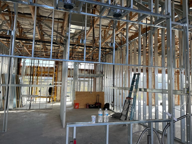 Interior of a construction site with metal stud framing, drywall partitions, wooden roof trusses, and construction equipment including a ladder and tool table
