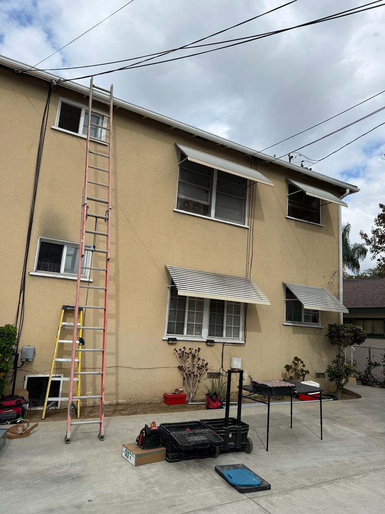 Pale yellow multi-story residential building with a metal ladder leaning against its side, concrete patio area with equipment below