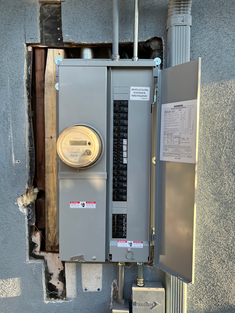 Outdoor electrical meter and breaker panel mounted on gray wall with doors open