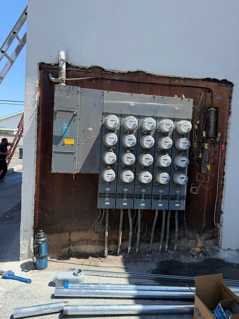 Exterior electrical meter panel mounted on a weathered brown metal box on a white wall with wiring and tools on the ground