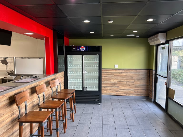 Interior of a modern fast-casual restaurant with red and lime green walls, wooden counter with bar seating, refrigerated beverage display, and contemporary lighting
