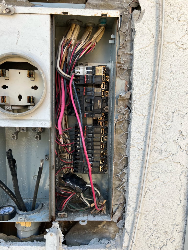 Open electrical fuse box with red and brown wires connected to multiple circuit breakers and terminals on a concrete wall