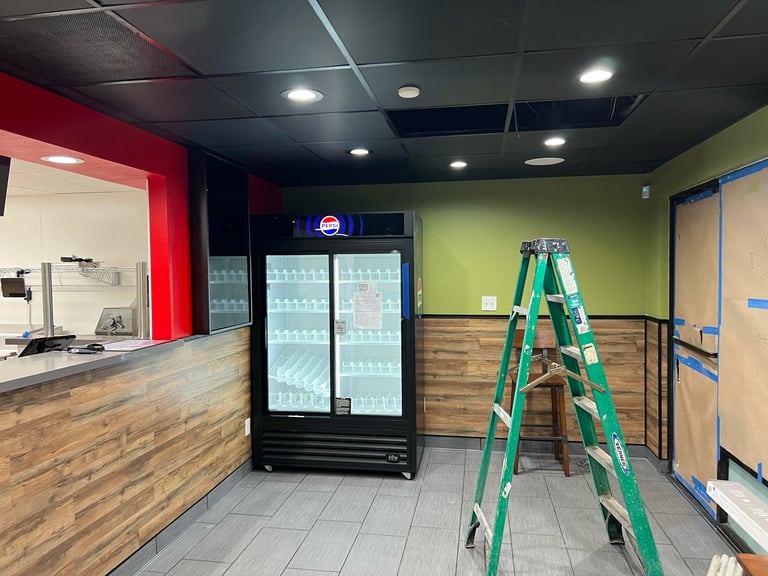 Interior of a small retail or food service space under construction with a refrigerated beverage cooler, green ladder, and colorful accent walls